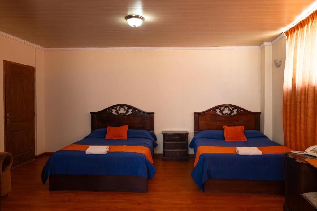 Habitación de hotel con dos camas de madera, cada una con una colcha azul y un cojín naranja. Entre las camas hay una mesita de noche de madera, y una puerta de madera al lado izquierdo.
