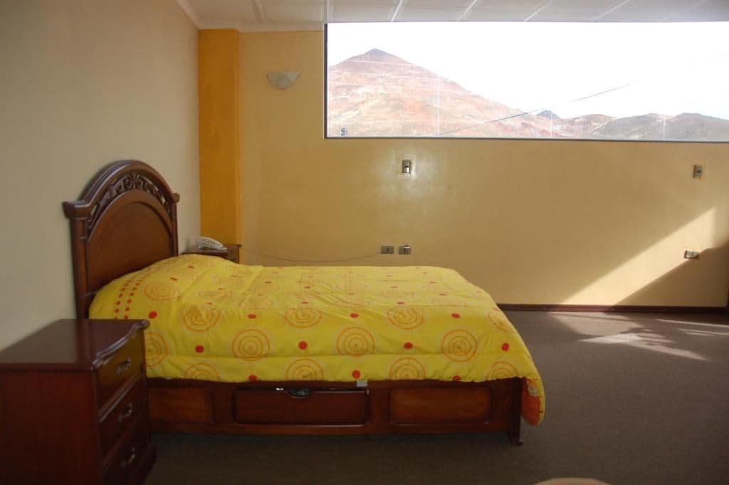 A bedroom with a bed, a wooden dresser, a window with a mountain view, and a wall-mounted light fixture.