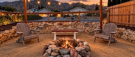 An outdoor seating area with a fire pit surrounded by rocks, two wooden chairs, and a wooden table.
