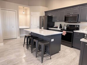 A modern kitchen with a central island, dark cabinetry, stainless steel appliances, and a light-colored floor. - Modern Retreat, Cozy, Calm & Perfectly Located (West Jordan)