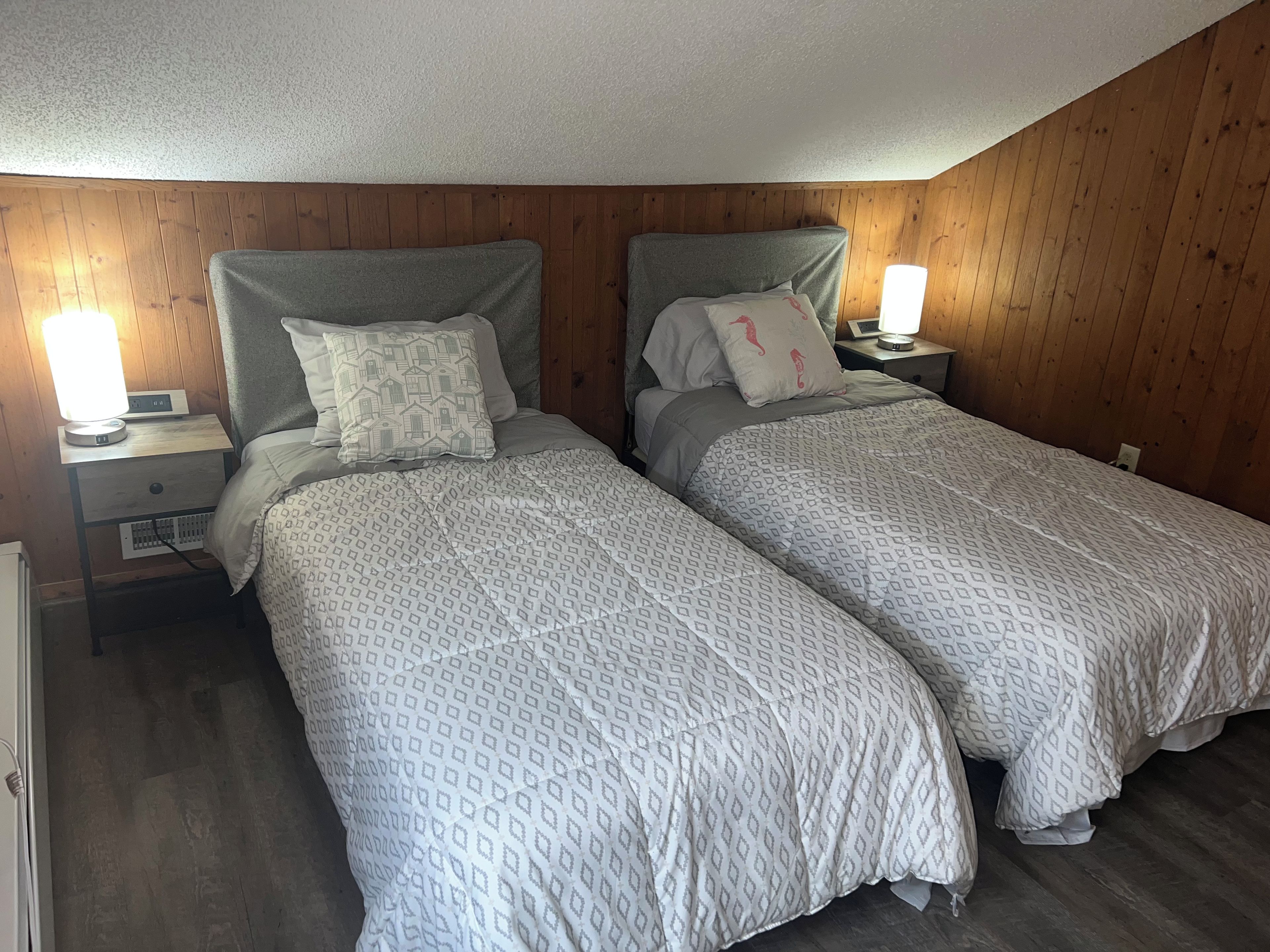 A bedroom with two single beds, wooden walls, a nightstand with a lamp, and a bedside table with a clock.