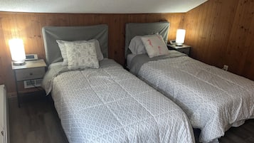 A bedroom with two single beds, wooden walls, a nightstand with a lamp, and a bedside table with a clock.