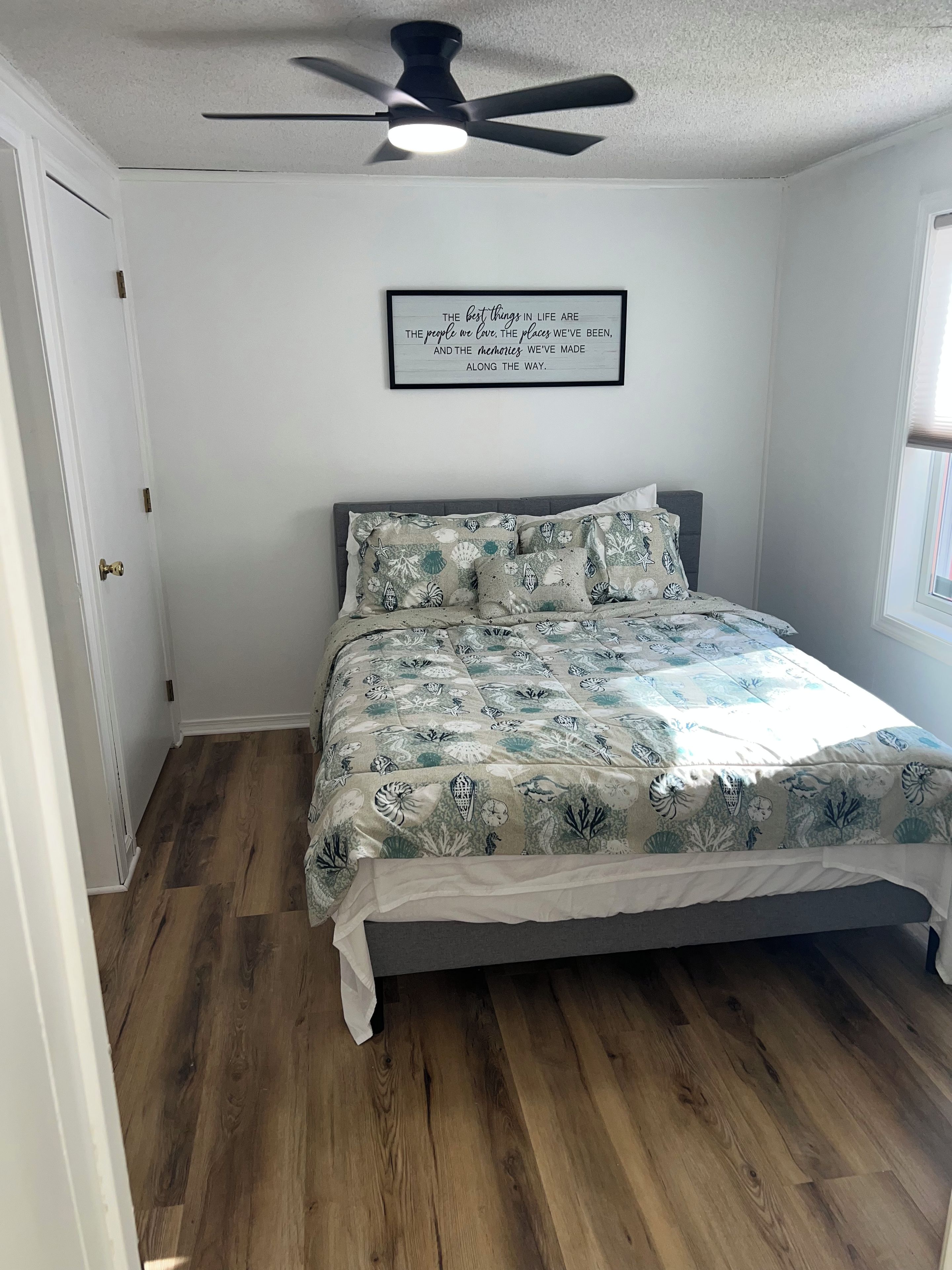 Un dormitorio con una cama, ventilador de techo, una cita enmarcada en la pared y una ventana.