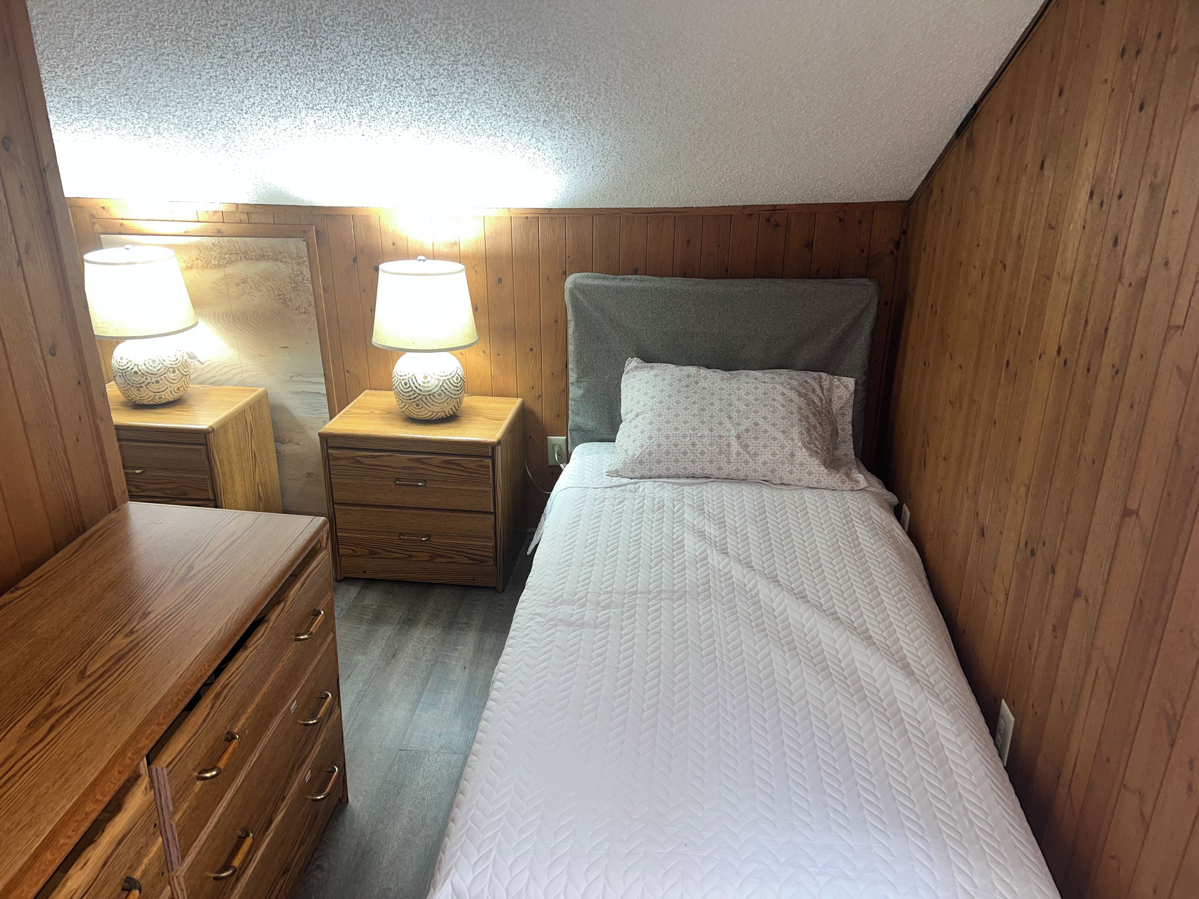 A small, cozy room with a single bed, a wooden nightstand with a lamp, and a dresser with drawers.