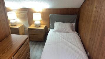 A small, cozy room with a single bed, a wooden nightstand with a lamp, and a dresser with drawers.