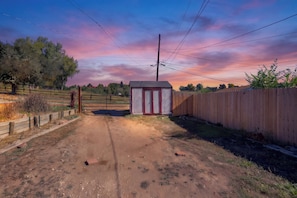 A dirt path leading to a red and white shed with a metal gate, surrounded by a wooden fence and trees. - Westside Bungalow | King Bed + Fenced Yard + Pets! (Colorado Springs)