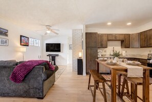 A modern living room with a grey sofa, a wooden dining table set for four, and a kitchen area with dark wooden cabinets and a white brick accent wall. - Westside Bungalow | King Bed + Fenced Yard + Pets! (Colorado Springs)