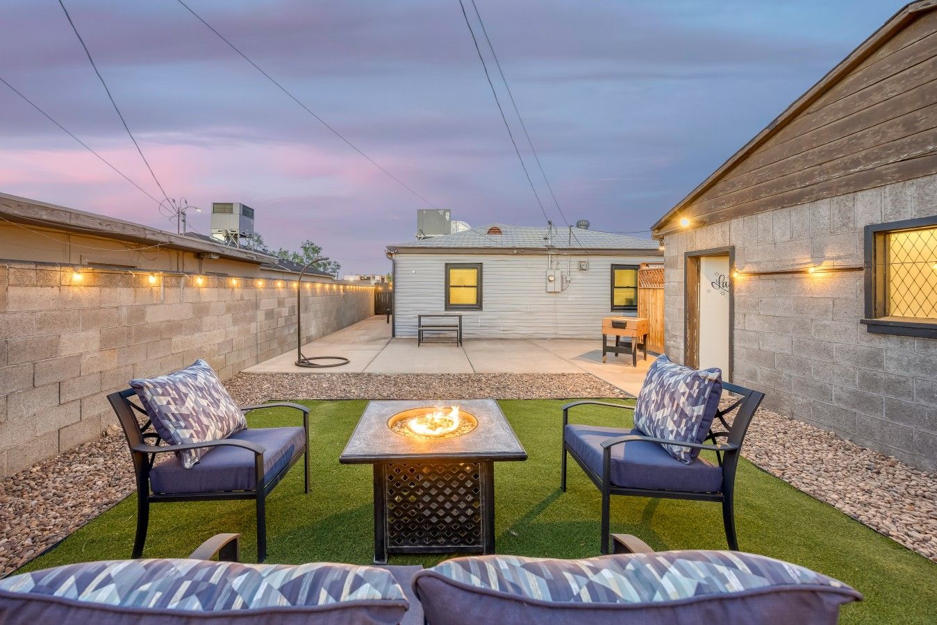 A backyard with a fire pit, two chairs, a table, and a small building with a window.