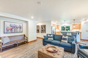 Ein modernes Wohnzimmer mit einer Couch, einer Bank und einem Couchtisch. Im Hintergrund befindet sich eine Küche mit Holzschränken und einem Fenster.