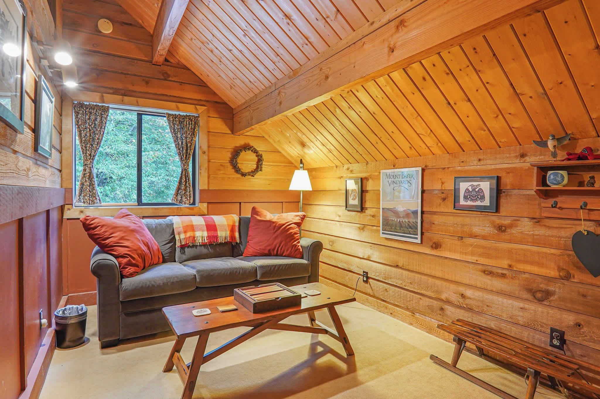 A cozy cabin interior with a wooden ceiling, a sofa with cushions, a small table, and a window with curtains.