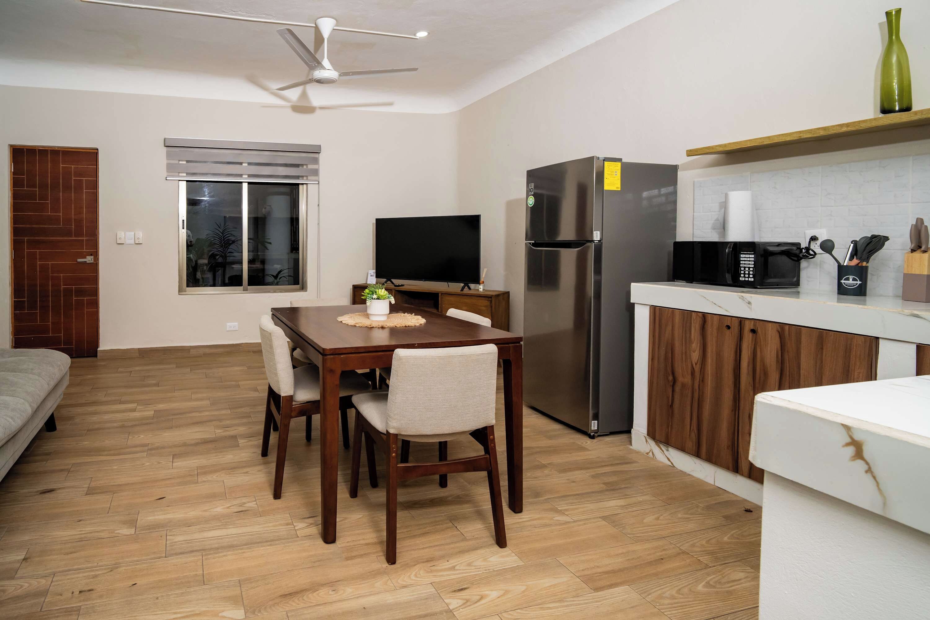 A compact living space with a dining area, a kitchen with a refrigerator and microwave, a television, and a ceiling fan.