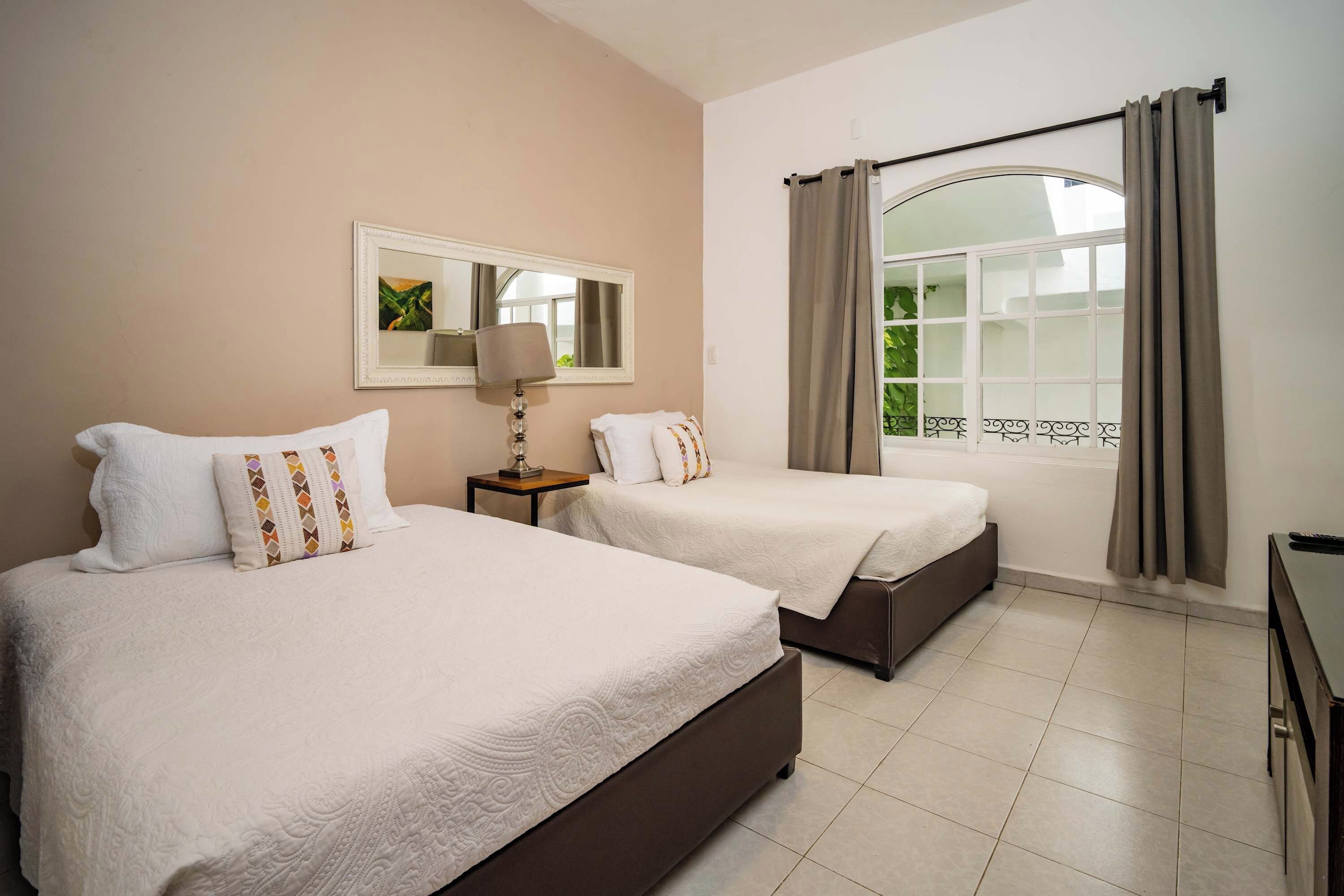 Une chambre d’hôtel avec deux lits, une table de chevet avec une lampe, un miroir et une fenêtre avec des rideaux.