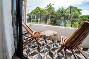 Ein Balkon mit zwei Holzstühlen, einem kleinen Tisch, einem gemusterten Boden und Blick auf Palmen und Grünpflanzen.
