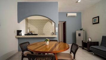 A compact living space with a dining area featuring a round wooden table and wicker chairs, a kitchenette with a coffee machine, and a grey sofa.