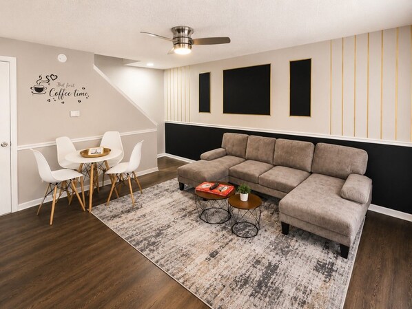 A modern living room with a sectional sofa, a dining area with white chairs, a coffee table, and a ceiling fan.