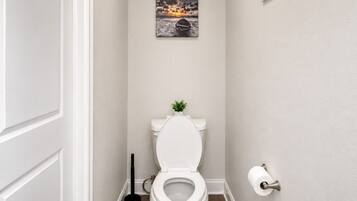 A bathroom with a white toilet, a small plant on the tank, a plunger, and a toilet paper holder.