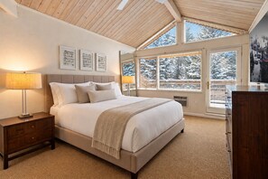 A bedroom with a large bed, bedside lamps, a wooden nightstand, a headboard, and a view of a snowy landscape through large windows. - Downtown Aspen | Pool & Hot Tub | Walk to Gondola by Aspen Vacation Rentals™ (Aspen)