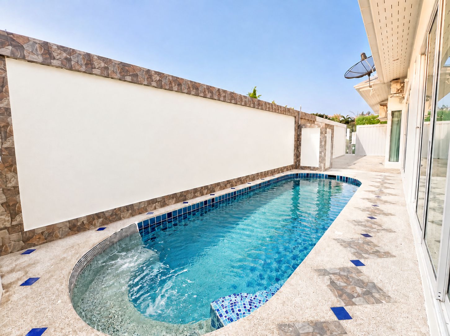 A swimming pool with a stone wall and tiled floor, surrounded by a building with a glass door.