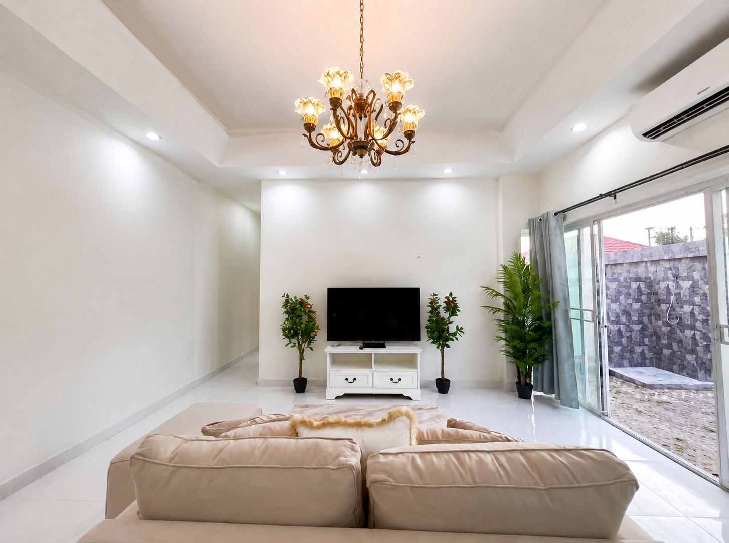 Un salon moderne avec un téléviseur à écran plat, un canapé beige, des plantes décoratives et une porte en verre donnant sur l’extérieur.