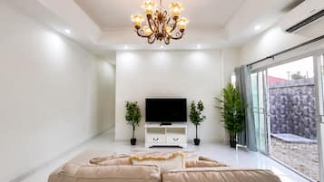 Un salon moderne avec un téléviseur à écran plat, un canapé beige, des plantes décoratives et une porte en verre donnant sur l’extérieur.