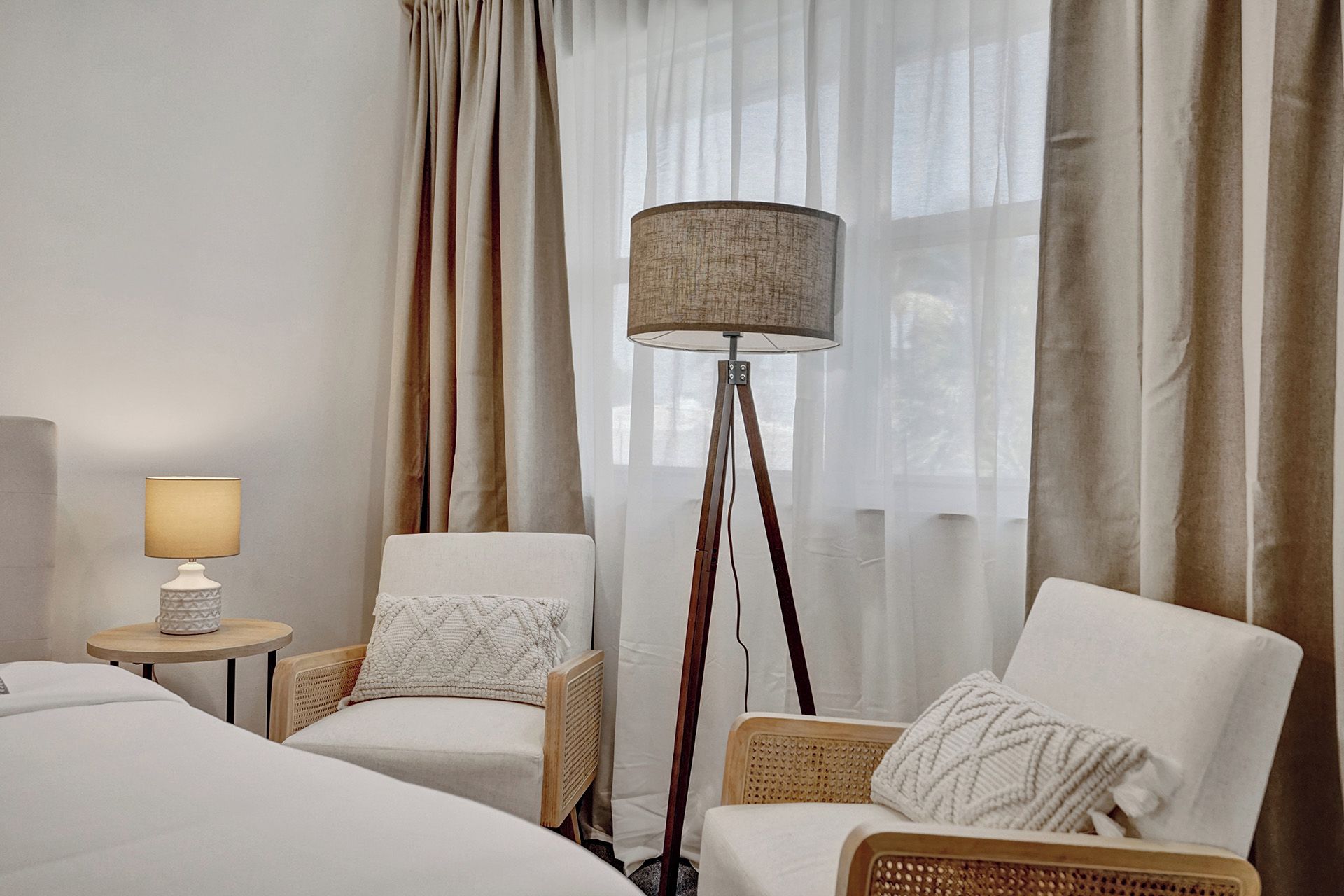 Ett hotellrum med en säng, två stolar, en golvlampa och tunna, genomskinliga gardiner.