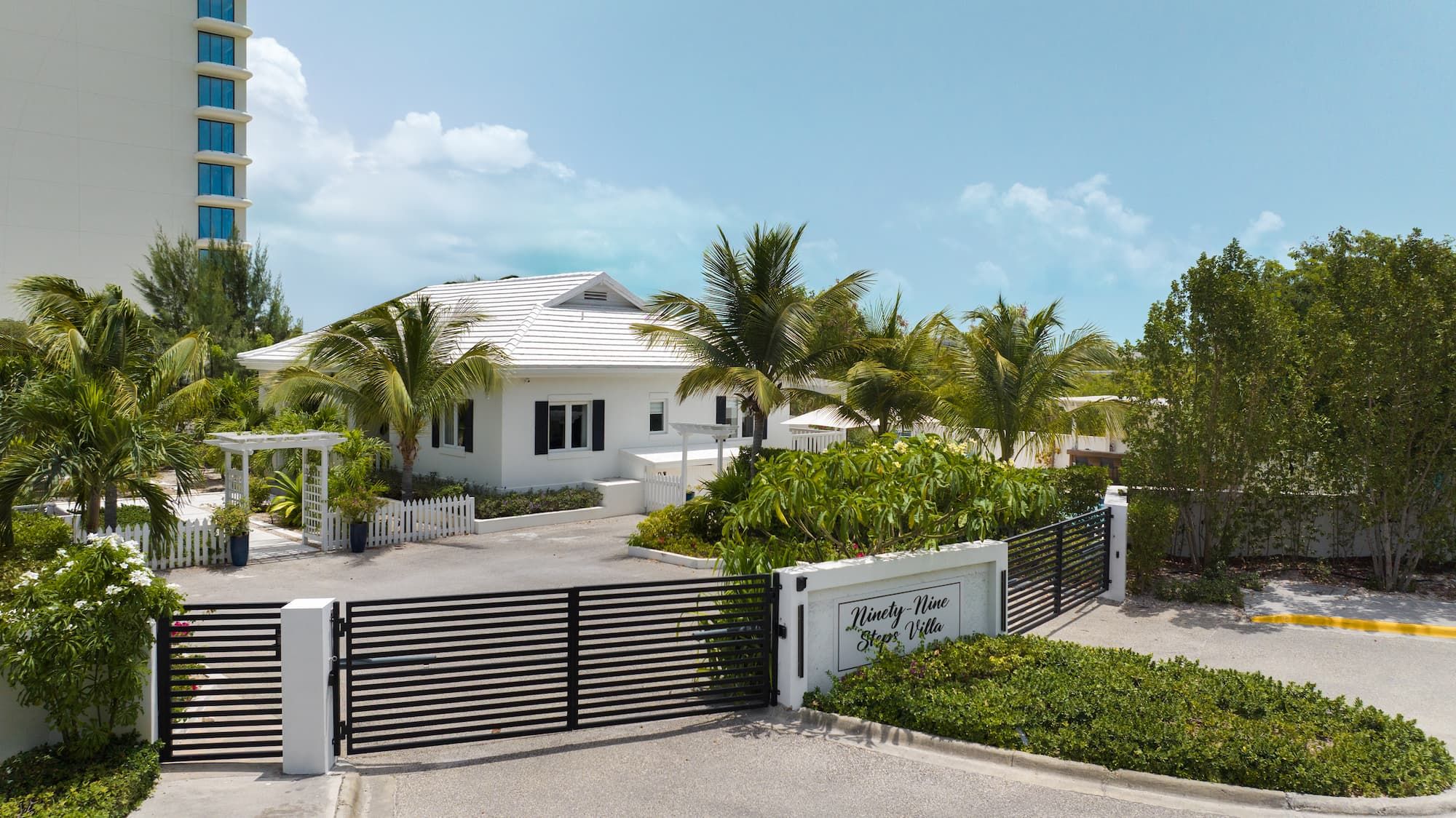 A white villa with a gated entrance, surrounded by lush greenery and palm trees.