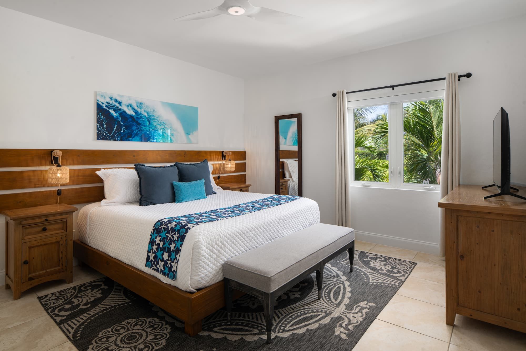 A bedroom with a bed, bedside tables, a bench, a TV stand, and a window with a view of greenery.