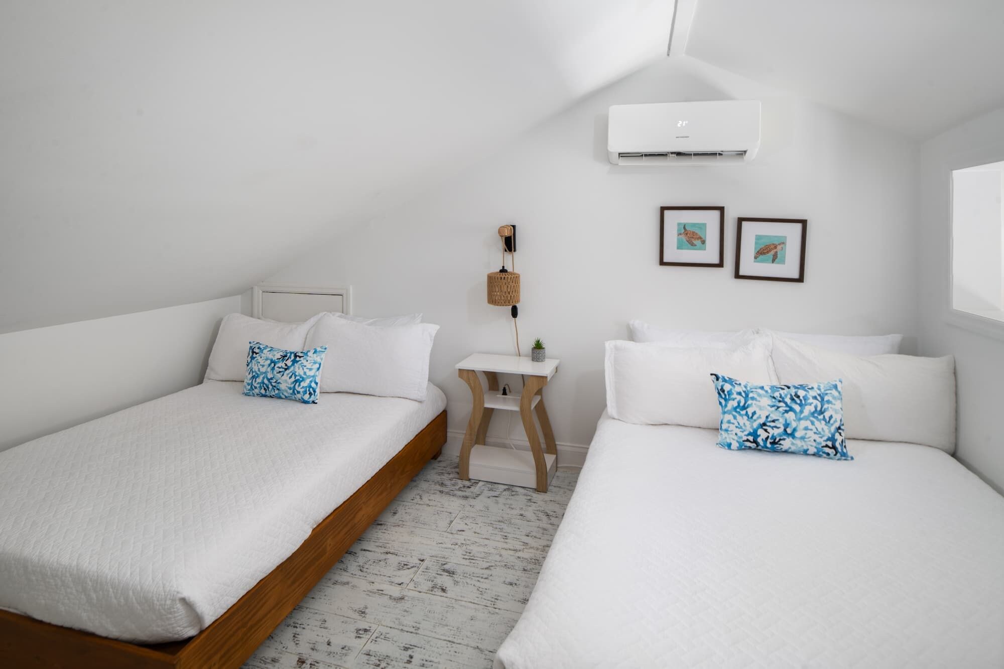 A bedroom with two beds, a small table, an air conditioning unit, and framed pictures on the wall.