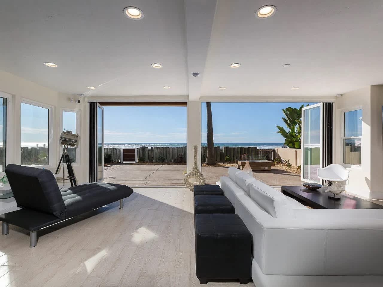 A modern living room with a large glass door leading to an outdoor terrace, a black lounge chair, a white sofa, and a tripod-mounted camera.