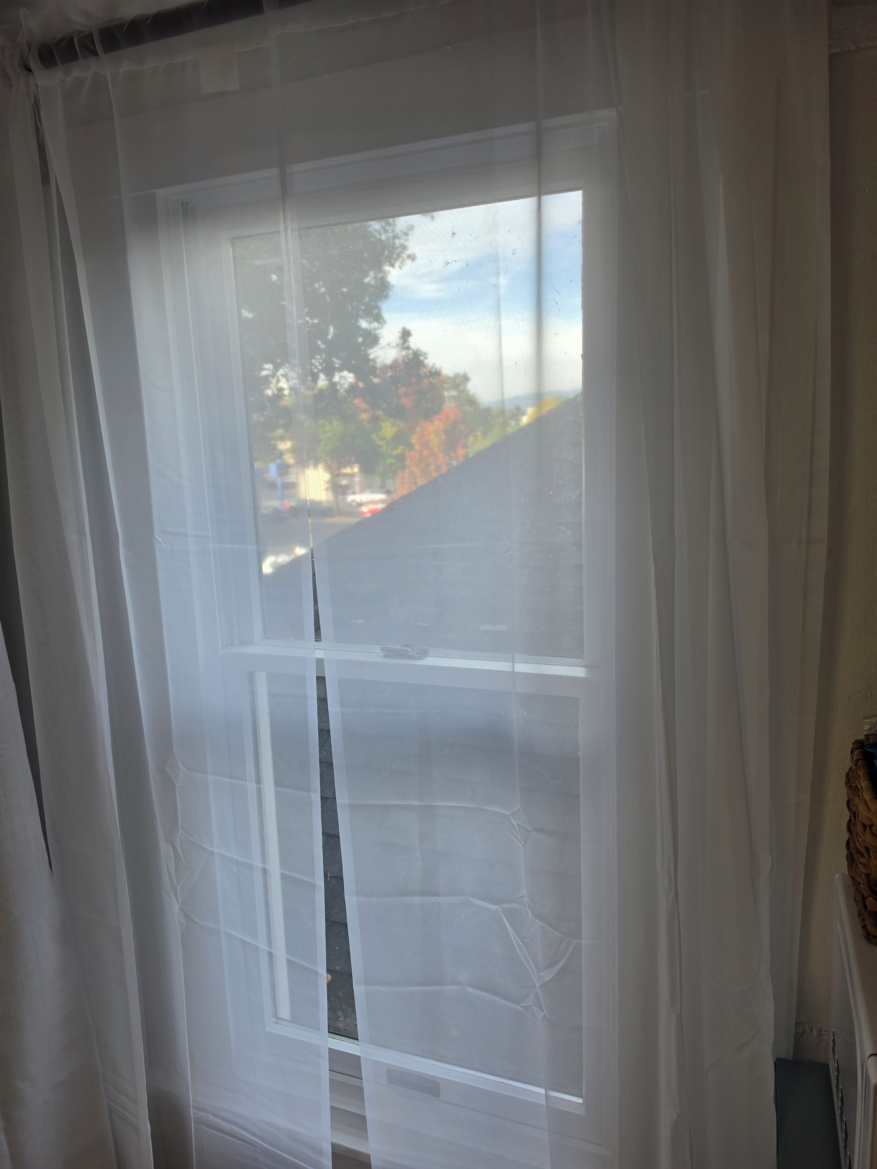 Una ventana con una cortina blanca translúcida entreabierta, que muestra vistas de árboles y un edificio.