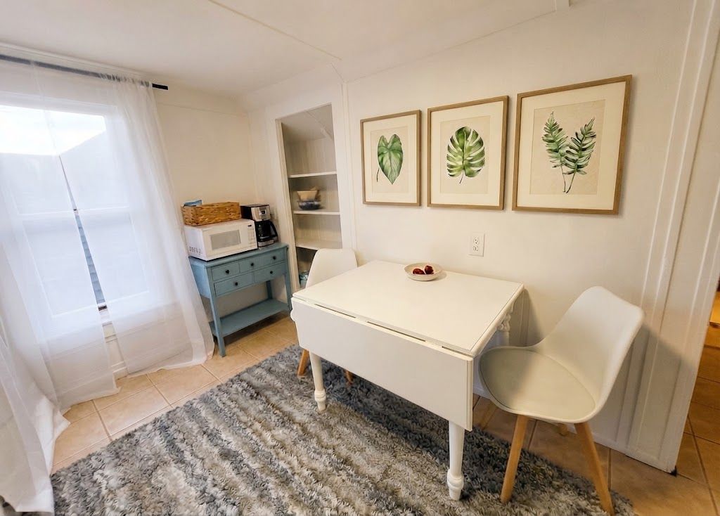 Een kleine, goed verlichte kamer met een witte tafel, twee stoelen, een witte magnetron, een blauwe kast en een raam met dunne gordijnen.