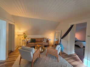 Een gezellige woonkamer met een bruine leren bank, een lichtgrijze fauteuil en een klein houten bijzettafeltje. Een witte vloerlamp en een potplant dragen bij aan de sfeer van de kamer.