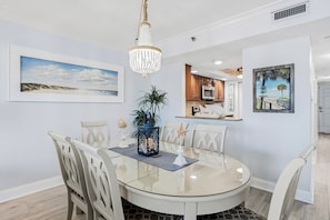 A dining area with a round glass table, white chairs, a chandelier, and a beach-themed painting. - Sandcastle North 302| 3br/2ba beach view (Indian Rocks Beach)