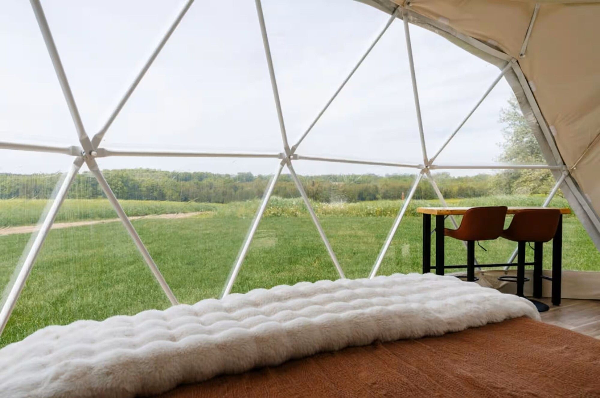 A tent with a view of a green field and trees. Inside, there is a bed with a white blanket and a table with chairs.