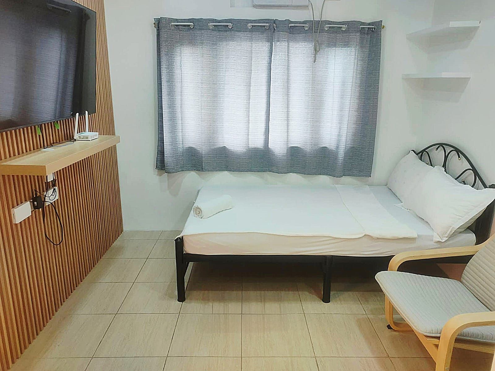 Ein Zimmer mit einem Bett, einem Sessel, einem Fernseher auf einer Holzablage und einem Fenster mit Vorhängen.