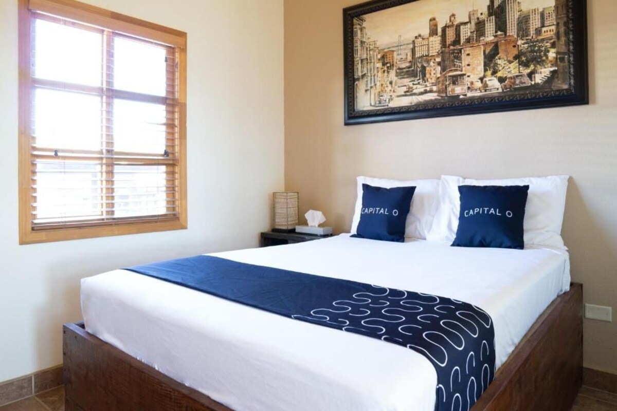 Une chambre d’hôtel avec un lit, une fenêtre avec des stores, une table de chevet, une lampe et un tableau représentant un paysage urbain accroché au mur.