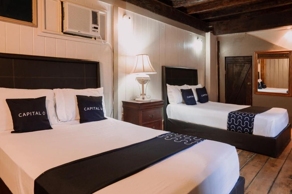 A hotel room with two beds, each with a black headboard and white bedding. There is a nightstand with a lamp between the beds.