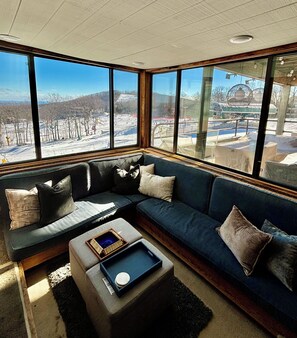 A corner seating area with a sofa and ottoman, a tray with a decorative item, and a view of a snowy landscape through large windows. - Dobie Ski-In Ski-Out + Mountain Views (Wintergreen Resort)