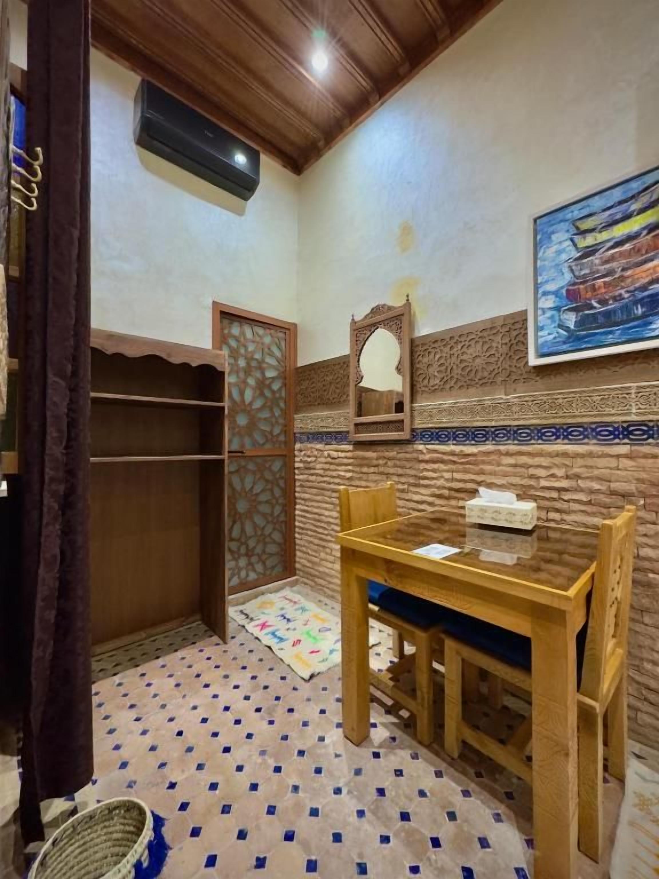 A room with a wooden ceiling, a patterned floor, a table with chairs, a mirror, and a wall-mounted air conditioner.