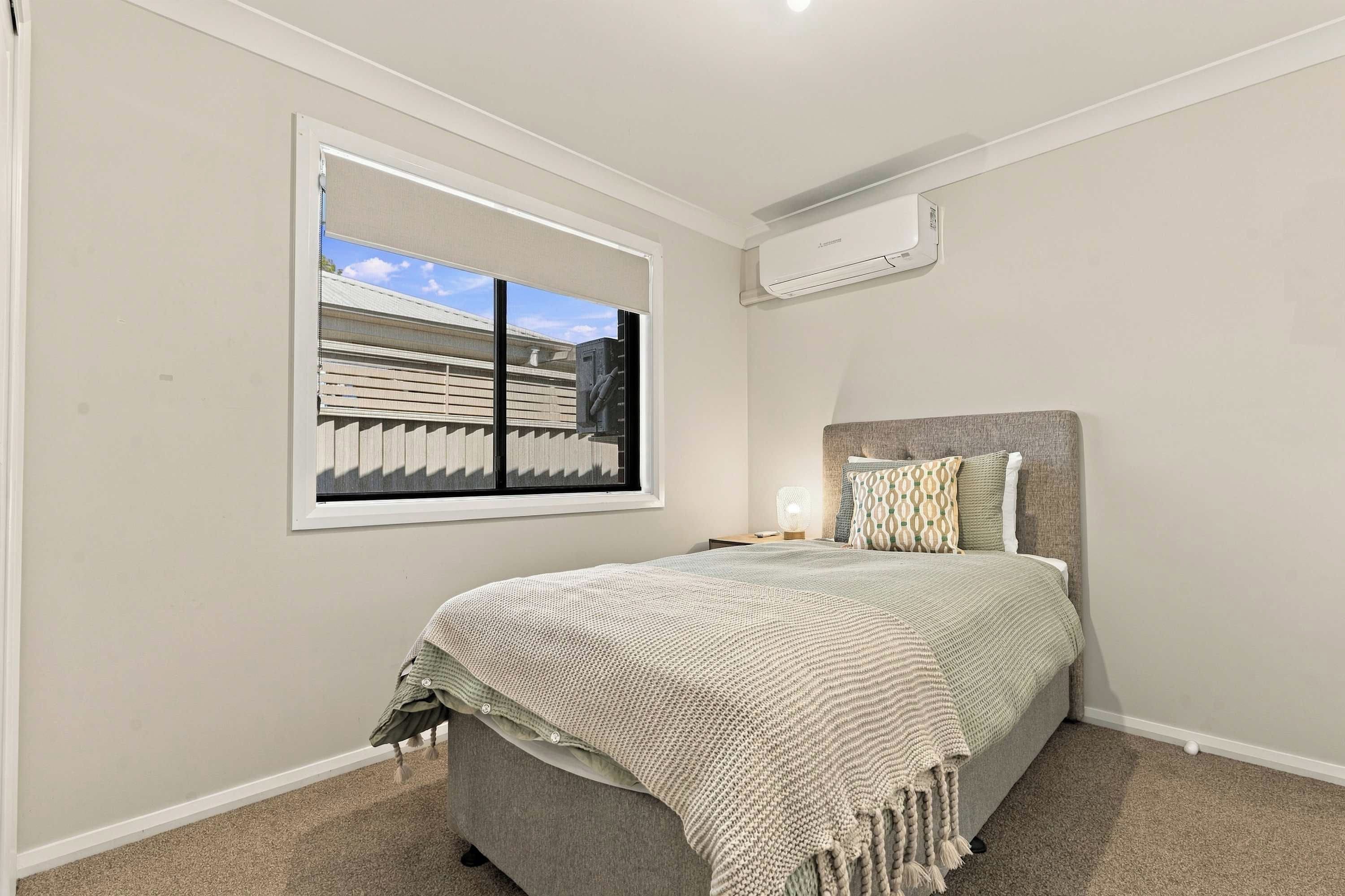 A bedroom with a bed, a window with blinds, an air conditioning unit, and a nightstand with a lamp.