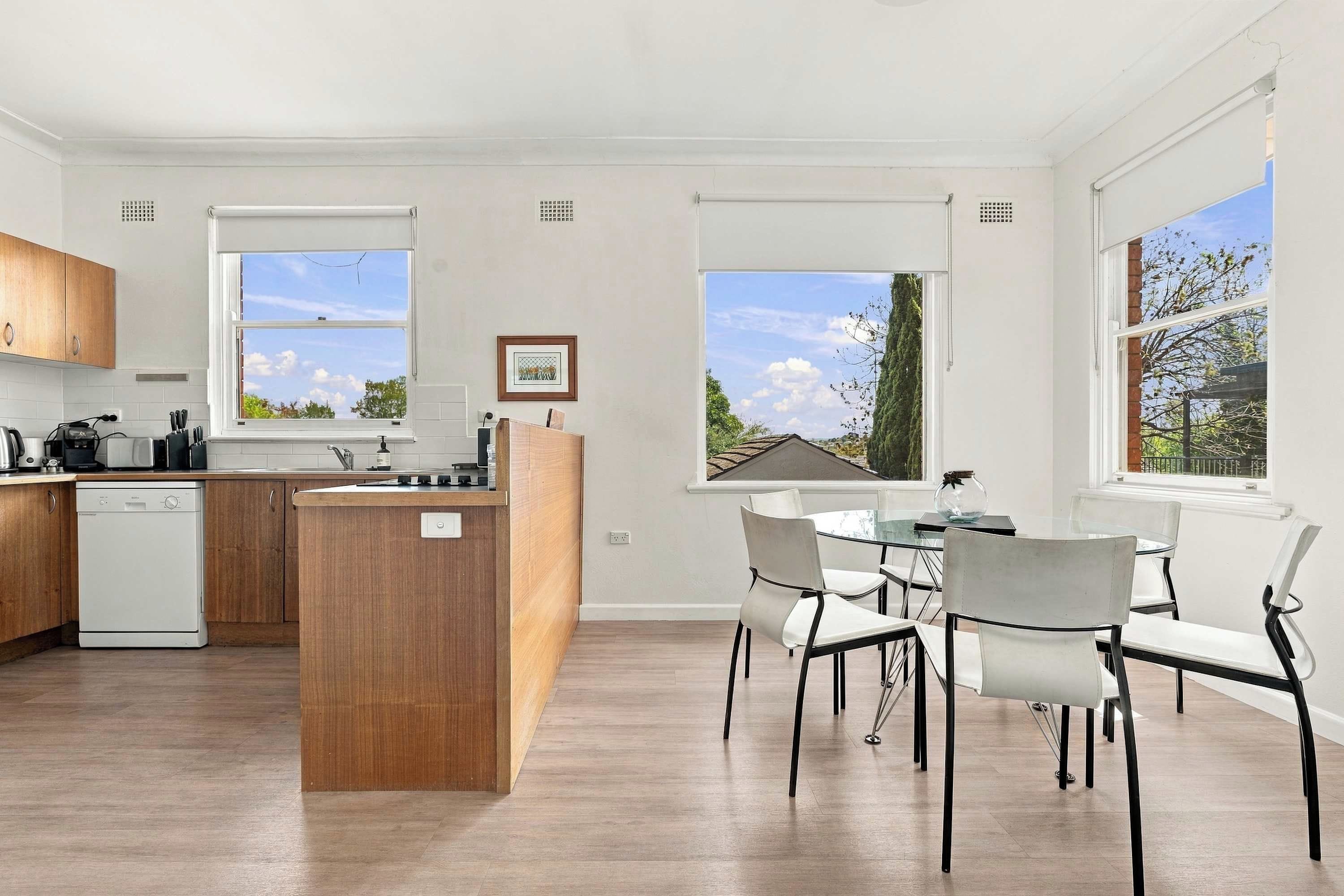A modern kitchen with a central island, dining area with chairs, and large windows offering views of the outdoors.