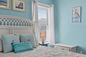 A bedroom with a bed, pillows, a nightstand, a lamp, a window with blinds, and a wall with a framed picture. - Oceanview Condo - Huge Deck Overlooking Ocean with Pool! (Carolina Beach)