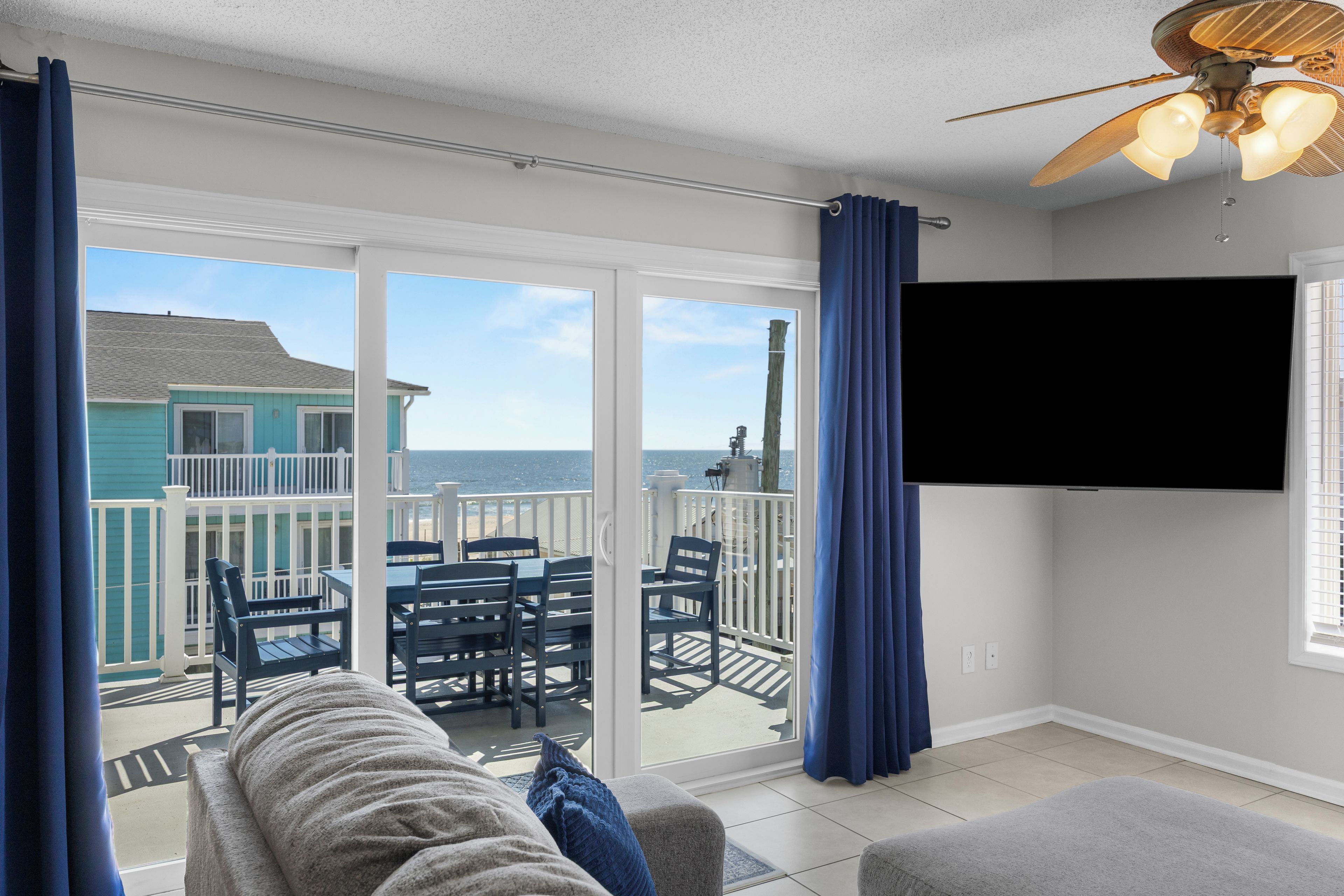 A room with a large sliding glass door leading to a balcony with a view of the ocean and a building. There is a ceiling fan, a flat-screen TV mounted on the wall, and a sofa with a blanket.
