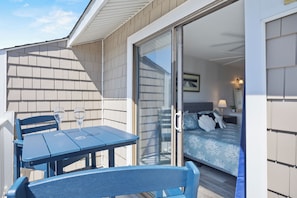 A balcony with a table and chairs, a bed with a blue bedspread, and a sliding glass door leading to a room with a nightstand and lamp. - Oceanview Condo - Huge Deck Overlooking Ocean with Pool! (Carolina Beach)