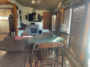 A cozy living room with a fireplace, a flat-screen TV, a dining table with chairs, and a large window with blinds. - The Captain at Secret Bay Hideaway (Private Oceanfront Cottage with Moorage) (Egmont)