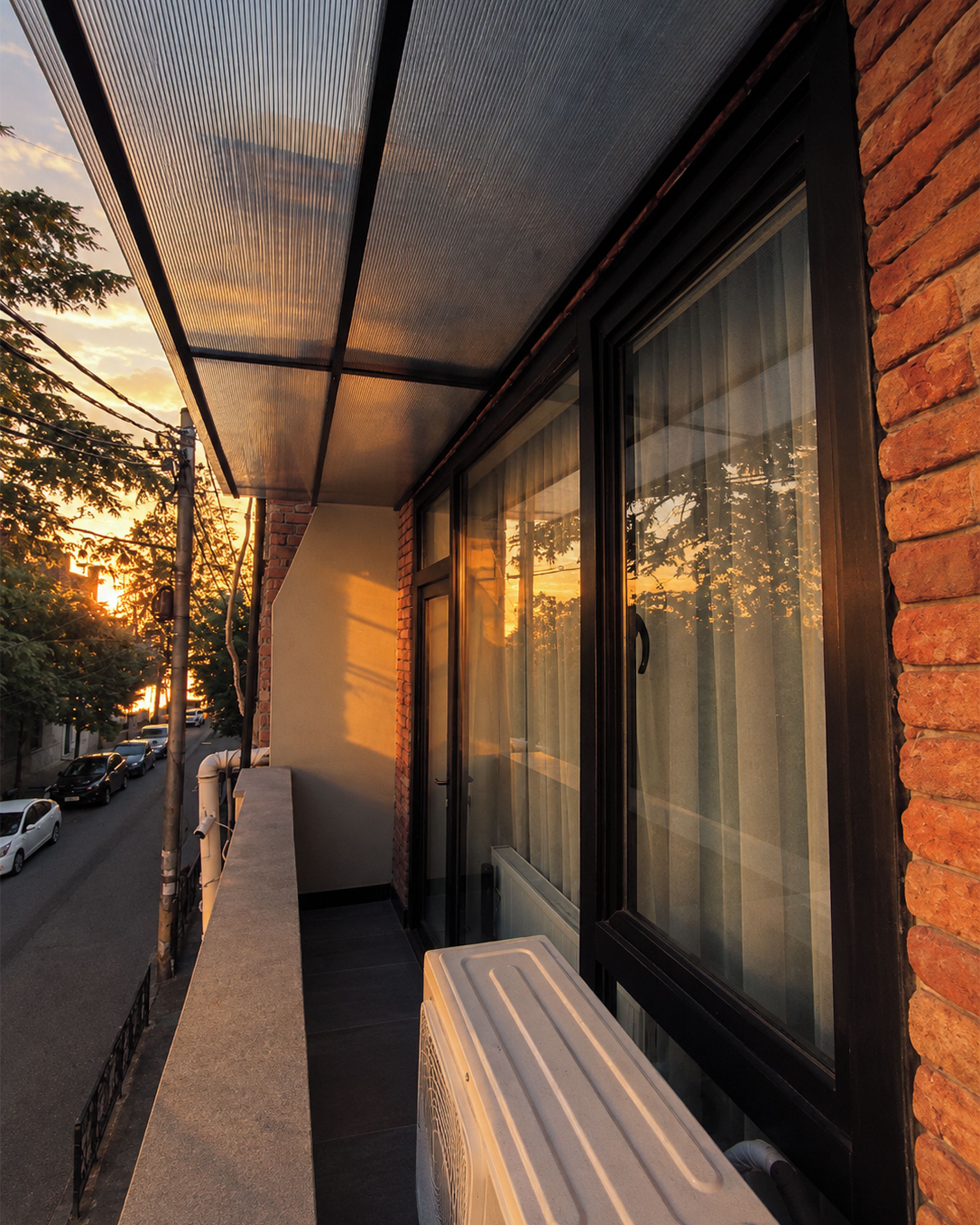 A covered outdoor area with a bench, a window with curtains, and a brick wall.