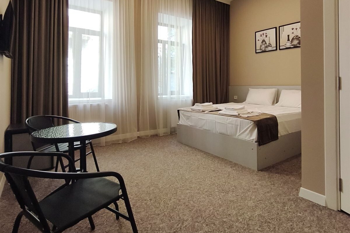 Quarto de hotel com cama, mesa pequena, duas cadeiras, janela com cortinas e TV fixada na parede.