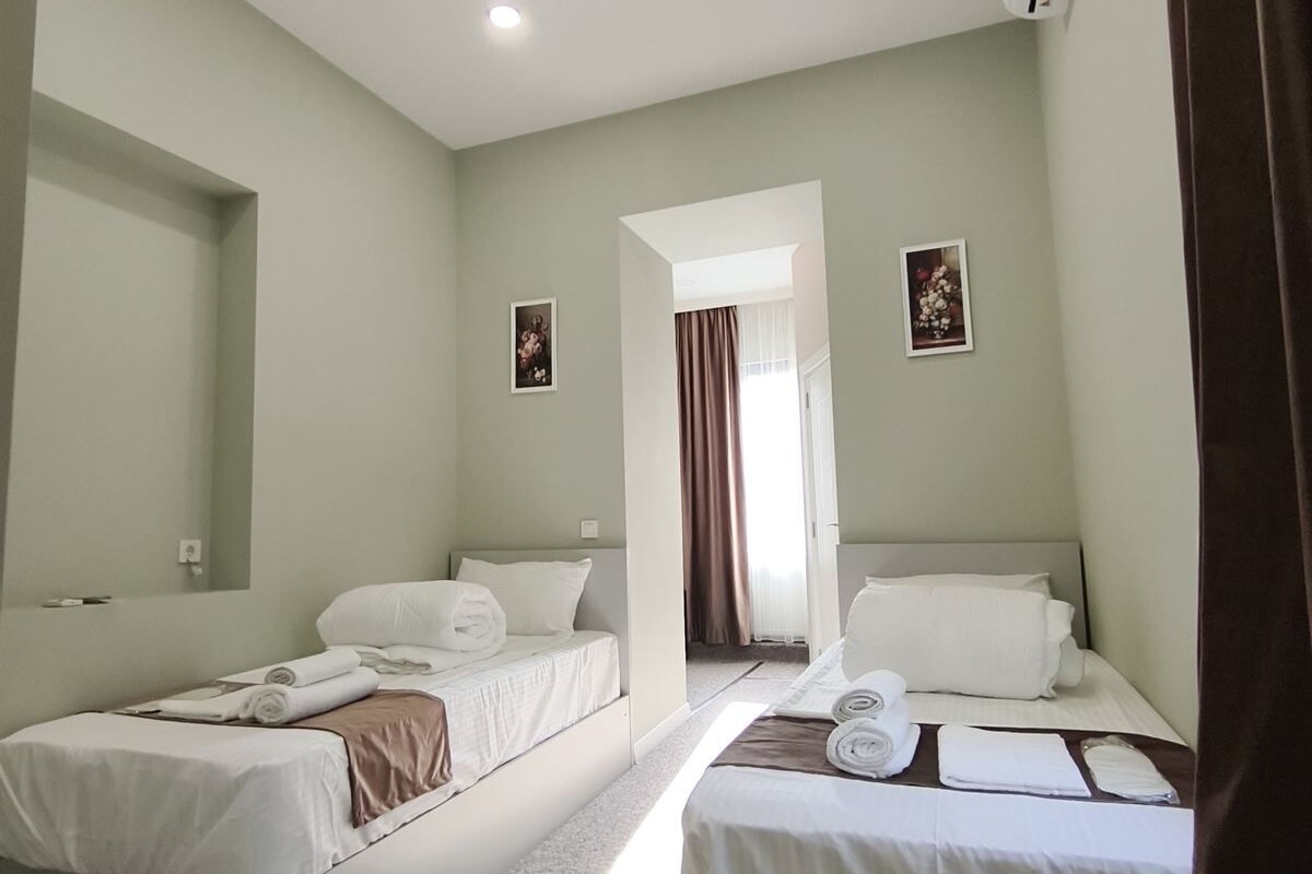 Quarto de hotel com duas camas de solteiro, uma luminária no teto, aparelho de ar condicionado e vista para o corredor.