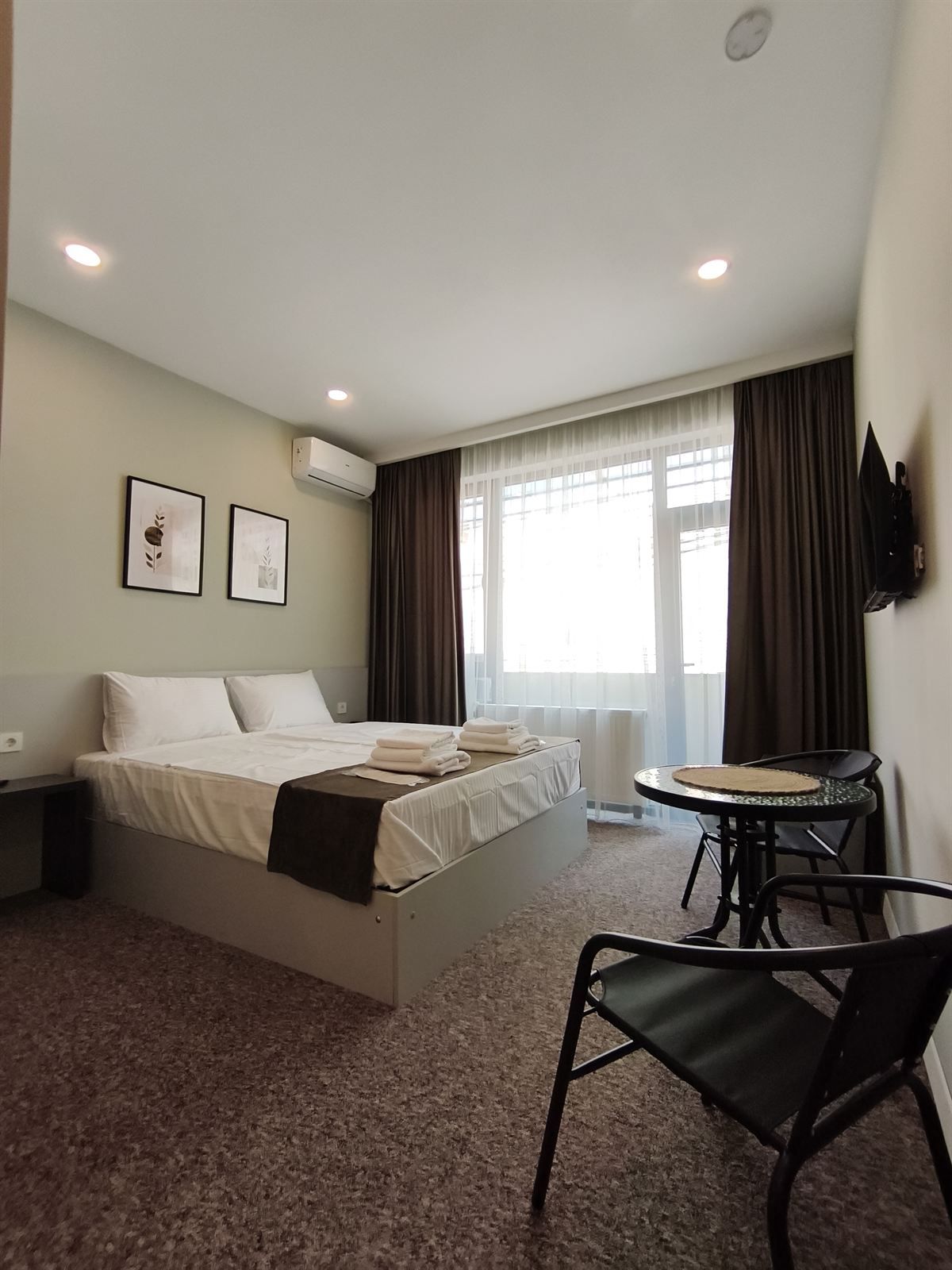 Quarto de hotel com uma cama, uma mesinha redonda, uma cadeira e uma televisão fixada na parede.