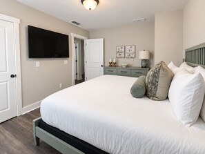 A bedroom with a large bed, a flat-screen TV mounted on the wall, a bedside table with a lamp, and a door leading to another room.